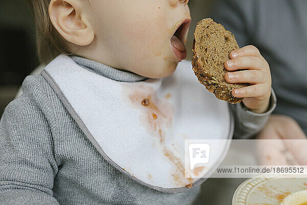 Baby boy with messy bib eating bread at home