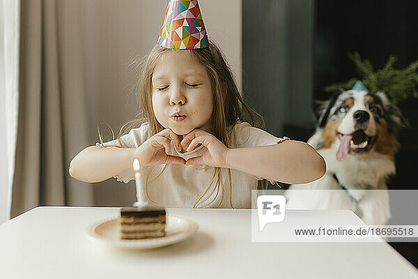 Girl making heart gesture and blowing candle on cake at home