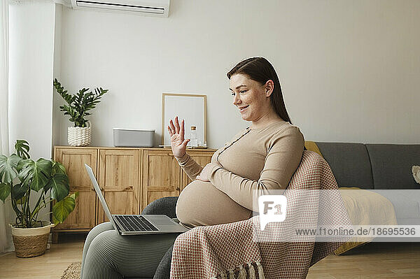 Smiling pregnant woman waving on video call through laptop at home