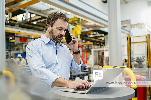 Manager talking on mobile phone using laptop at factory