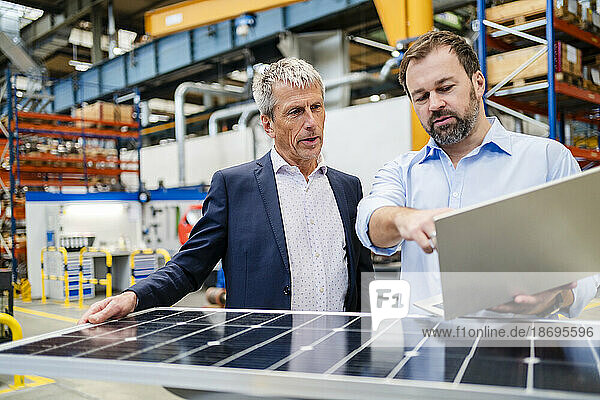 Manager holding laptop by businessman holding solar panel at factory
