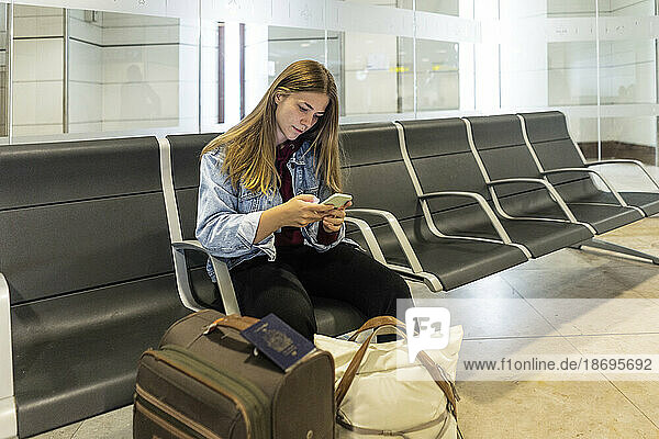 Young woman using smart phone sitting on seat at airport lobby