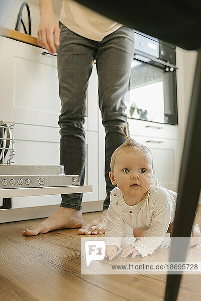Cute baby girl crawling on floor by father loading utensils in dishwasher at home