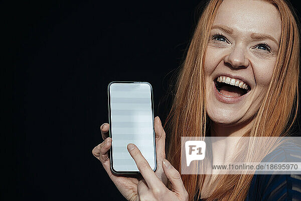 Junge Frau lacht und hält Smartphone vor schwarzem Hintergrund