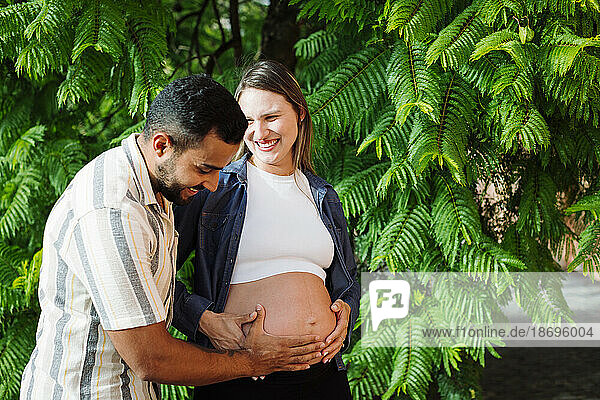 Smiling man touching belly of pregnant woman in front of tree