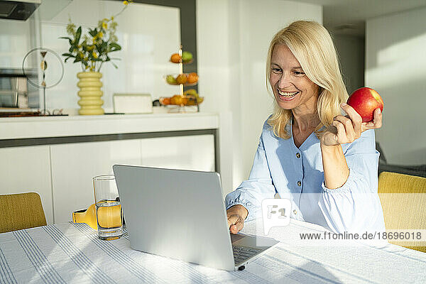 Happy blond freelancer holding apple and doing video call through laptop at home office