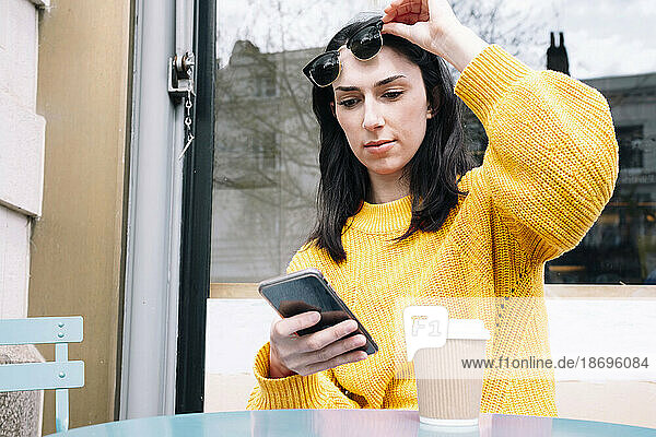 Woman in yellow sweater lifting sunglasses and using smartphone at outdoor cafe