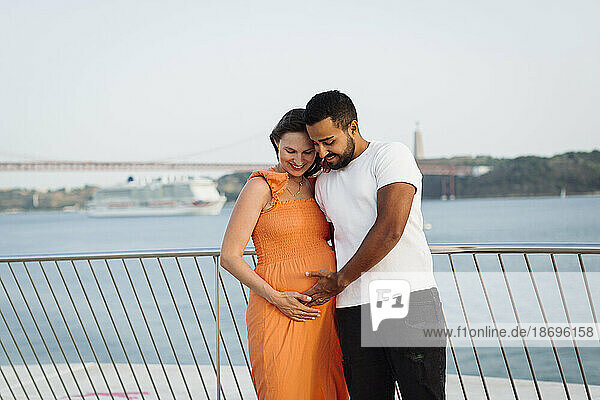 Man touching belly of pregnant woman standing in front of railing