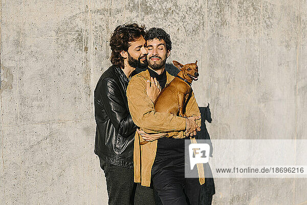 Gay man embracing boyfriend carrying dog in front of concrete wall