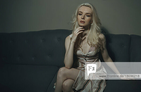 Blond woman with hand on chin sitting on sofa in darkroom