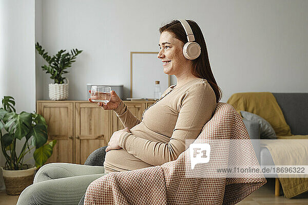 Smiling pregnant woman listening to music holding glass of water sitting at home