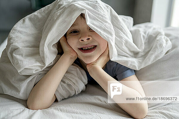Boy lying under blanket on bed at home