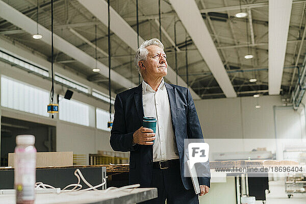 Thoughtful businessman holding reusable cup in factory