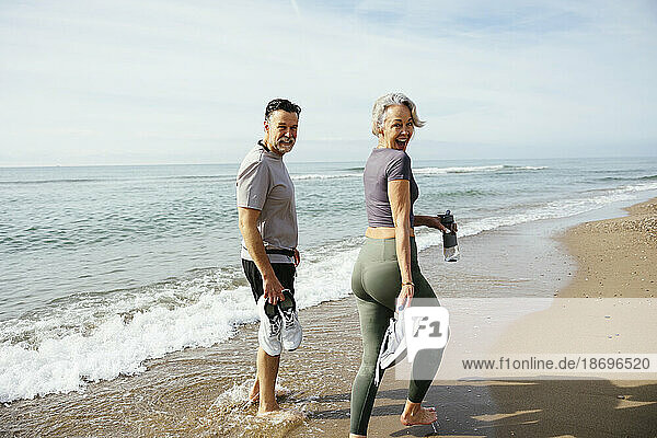 Mature couple holding sports shoes walking near shore at sunny day