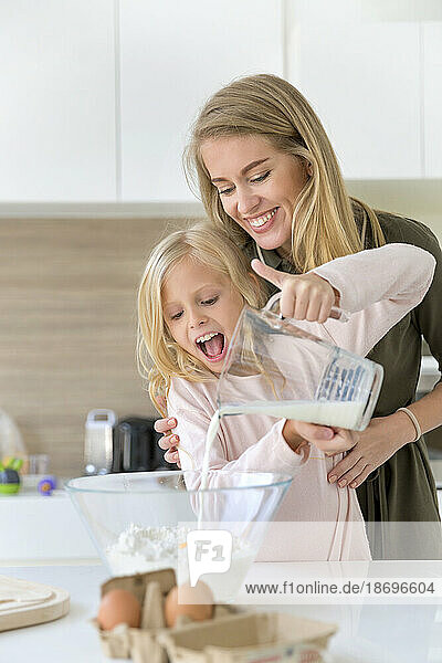 Lächelnde Frau hilft ihrer Tochter  zu Hause Milch in Mehl zu gießen