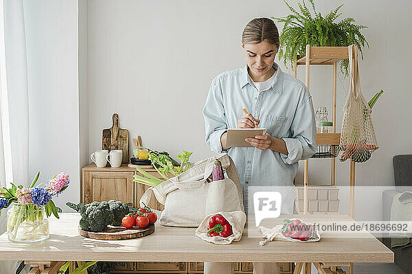 Woman preparing shopping list with groceries on table at home