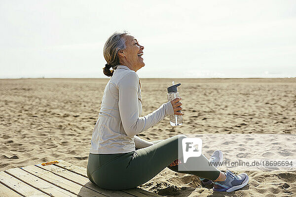 Woman with water bottle sitting and laughing at beach