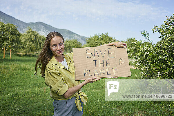 Lächelnde Frau zeigt den Text „Save The Planet“ auf einem im Garten ausgeschnittenen Karton