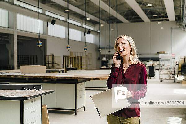 Glückliche Geschäftsfrau  die in der Fabrik mit einem Smartphone spricht und eine Datei in der Hand hält