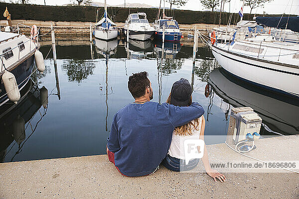 Man with arm around girlfriend sitting on jetty at harbor