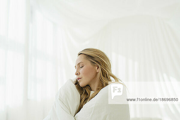 Woman with eyes closed wrapped in white blanket by translucent curtain