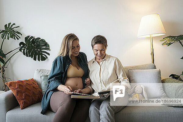 Mother and pregnant daughter sharing photo album sitting on sofa at home