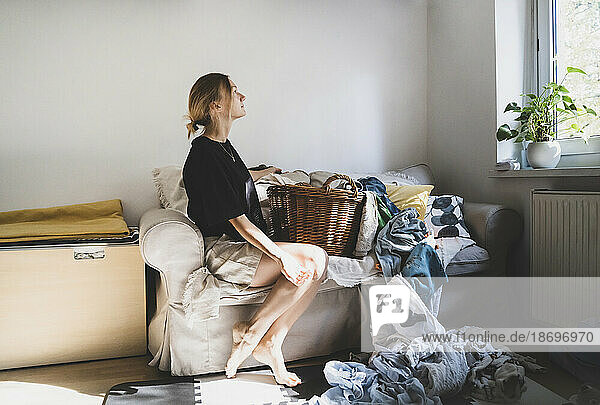 Thoughtful woman sitting on sofa with cluttered laundry at home