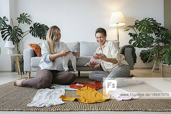 Happy mother and pregnant daughter looking at baby clothes sitting at home