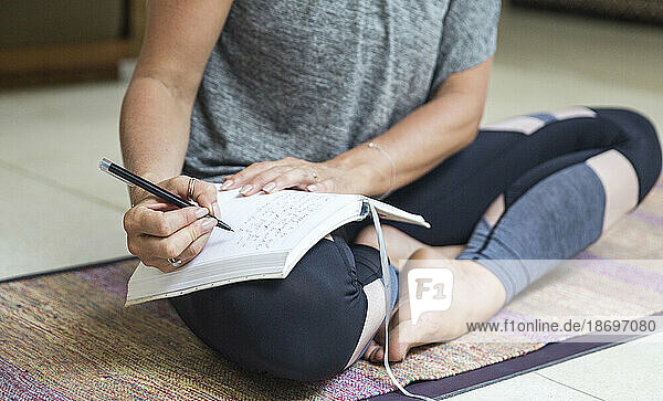 Frau schreibt Tagebuch und sitzt zu Hause mit gekreuzten Beinen auf einer Trainingsmatte