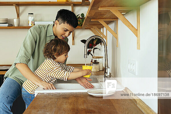 Son filling water in glass through faucet by mother in kitchen at home