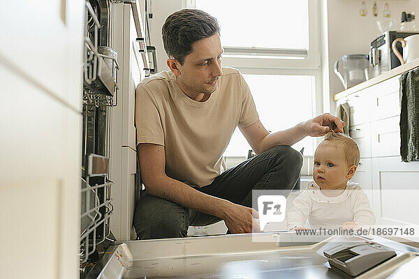 Cute baby girl and father loading utensils in dishwasher in kitchen at home