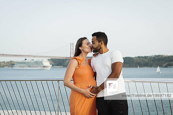 Schwangere Frau küsst Mann vor Geländer