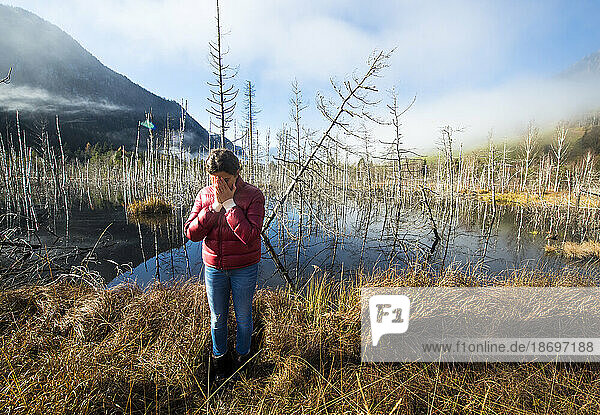 Depressed woman standing on dried grass in front of lake