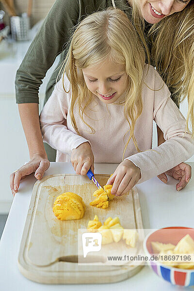 Smiling girl learning to chop pineapples on cutting board by mother at home