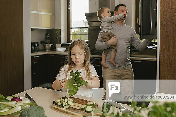 Girl holding Bok Choy with father carrying son in background at home