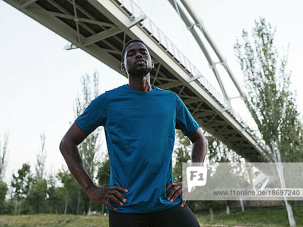 Confident athlete with arms akimbo standing under bridge