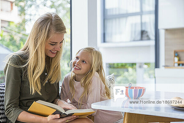 Lächelndes Mädchen schaut Mutter zu  wie sie zu Hause in der Küche ein Buch liest