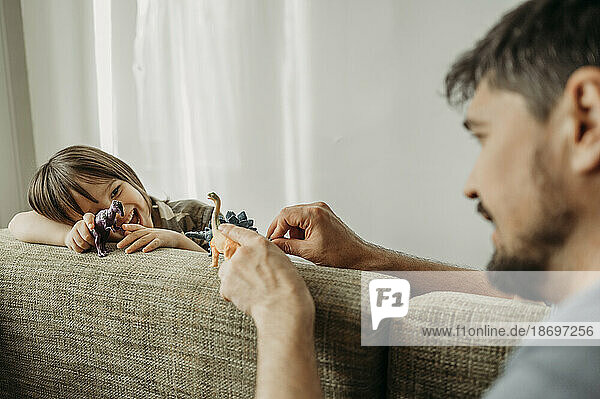 Father and son playing with toy dinosaurs on sofa at home