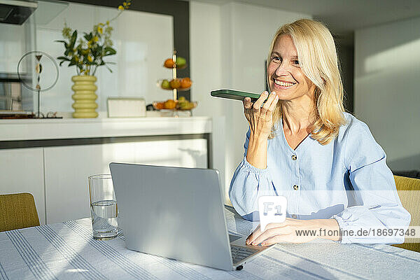 Smiling woman talking on speaker phone with laptop at desk in home office