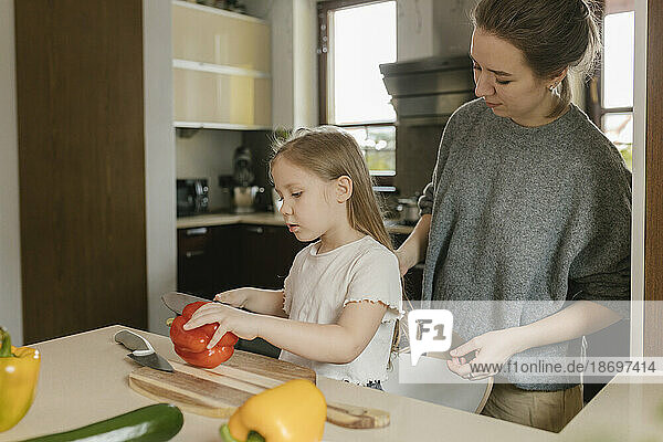 Mother with daughter cutting red bell pepper in kitchen at home