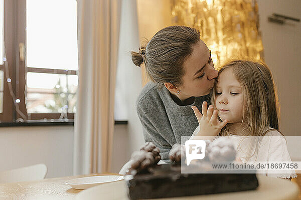 Mother kissing daughter by chocolate cake at table
