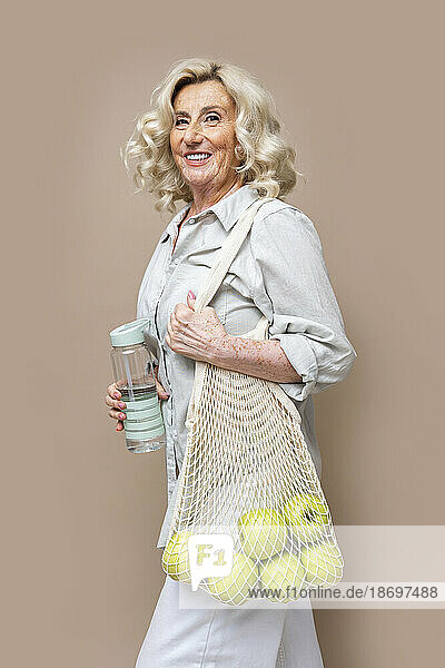 Smiling senior businesswoman with apples in mesh bag against beige background