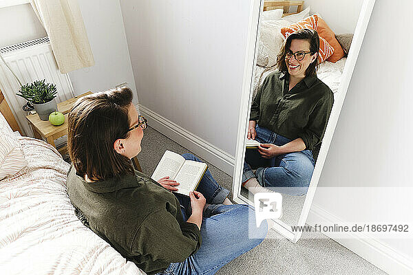 Happy woman sitting with book near mirror at home