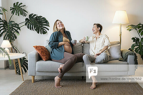 Happy pregnant daughter having coffee with mother sitting on sofa at home