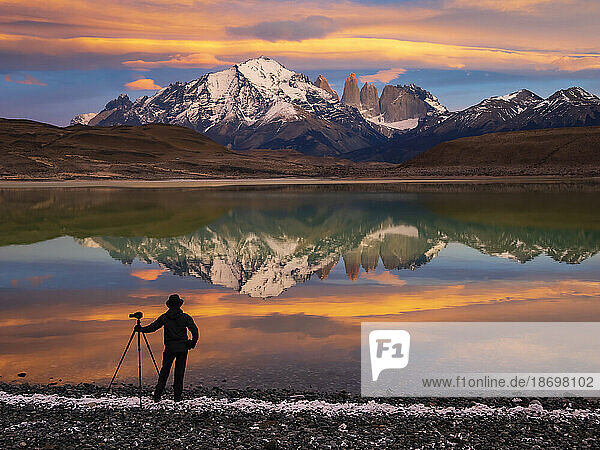View taken from behind of a silhouette of a photographer with a camera tripod standing at the water's edge at Lago Azul at sunrise tracking pumas; Torres del Paine National Park  Patagonia  Chile
