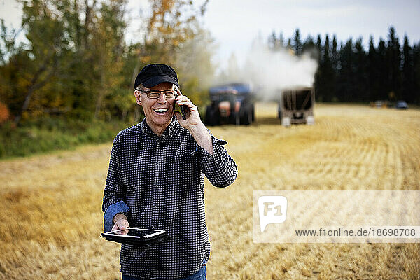 Farmer using a tablet to manage his harvest and talking on his cellphone with harvesting equipment working in the background; Alcomdale  Alberta  Canada