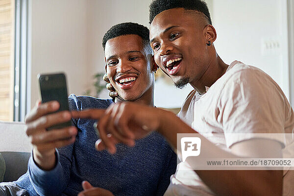 Happy young gay male couple using smart phone at home