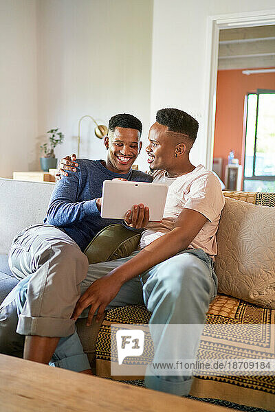 Happy young gay male couple using digital tablet on living room sofa