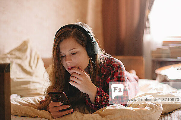 Young woman with headphones using smart phone lying on bed at home
