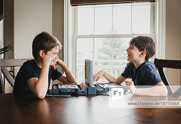 Two boys smiling as they play a board game together at a wooden table.